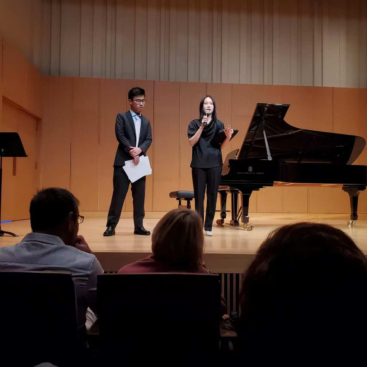 Yi-Hui Chen speaking into a microphone on stage beside a student in front of a grand piano