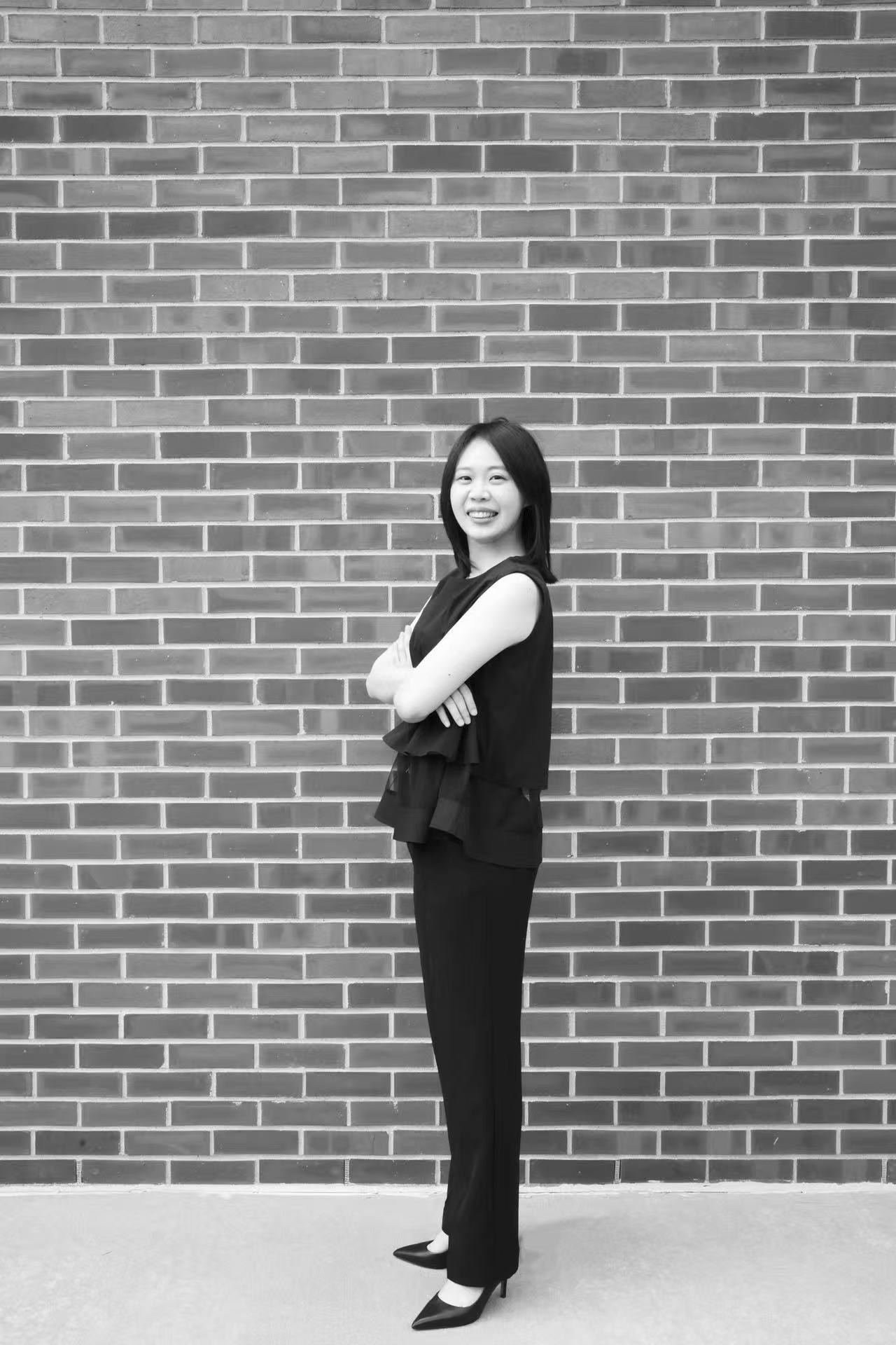 Black-and-white full-length portrait of Yi-Hui Chen smiling in front of a brick wall