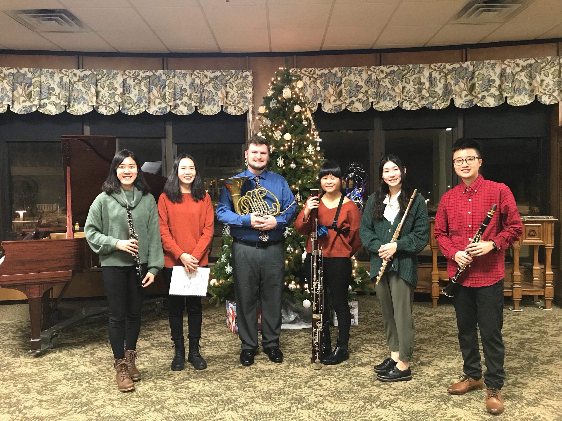 Yi-Hui Chen with a wind ensemble holding oboe, horn, bassoon, flute, and clarinet beside a decorated Christmas tree
